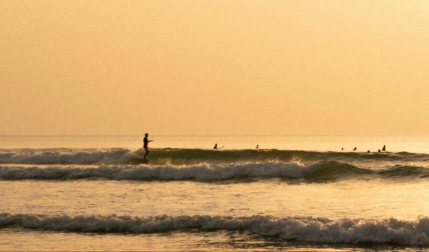 Surfers on a beach at sunset with a warm orange sky.